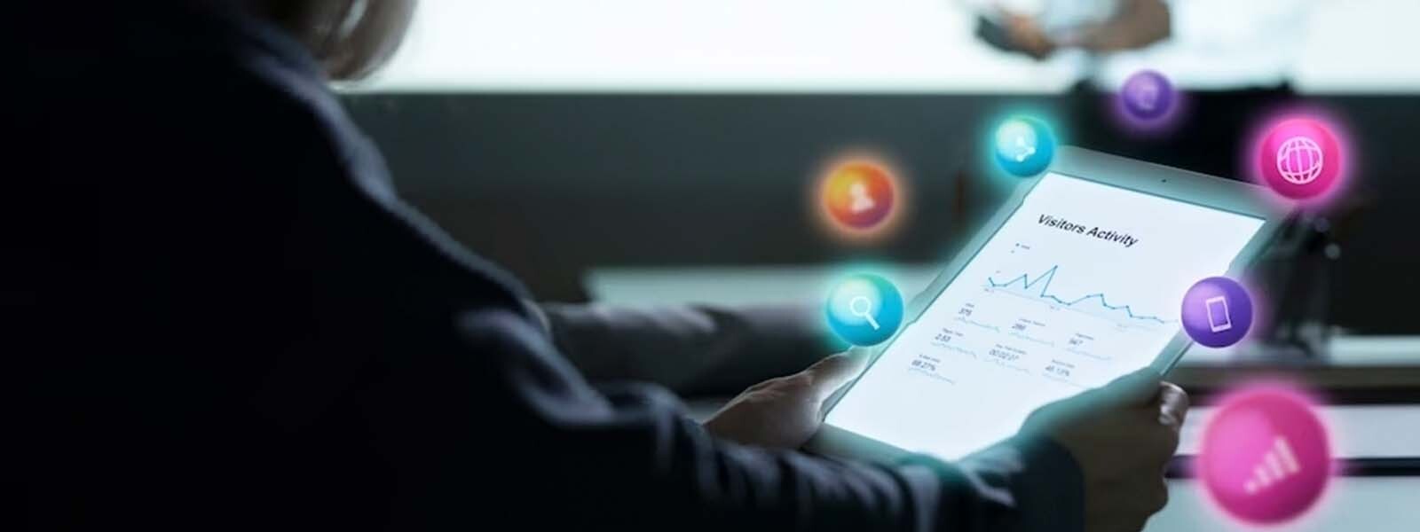 Persona sostiene una tablet en sus manos, dentro de la cual se visualiza un panel estadístico y varios iconos de marketing animados salen arriba de la tablet.